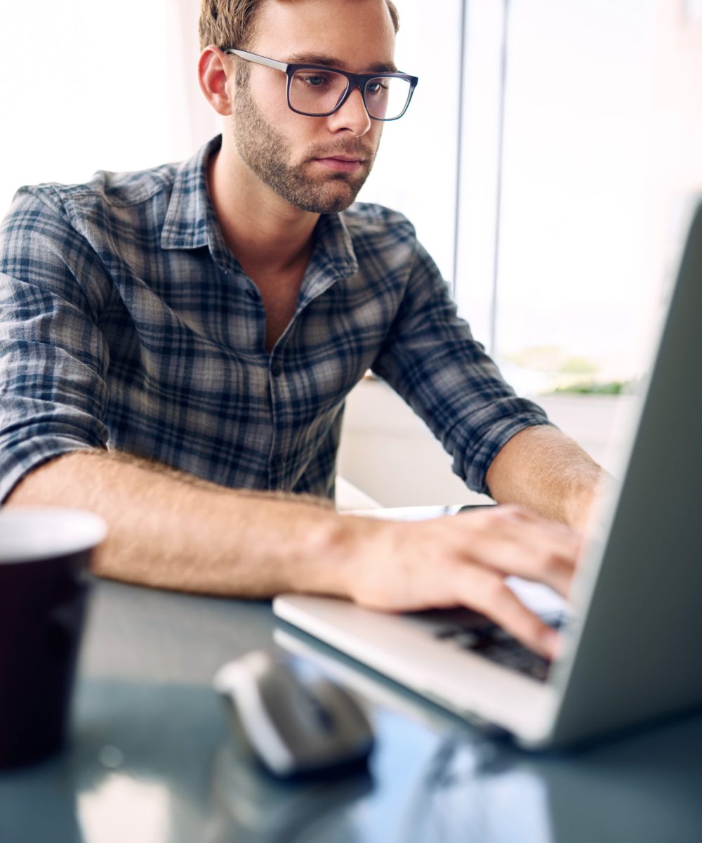Portrait image of a young businessman typing on his laptop while wearing a check shirt and glasses, with a cup of coffee on the ready