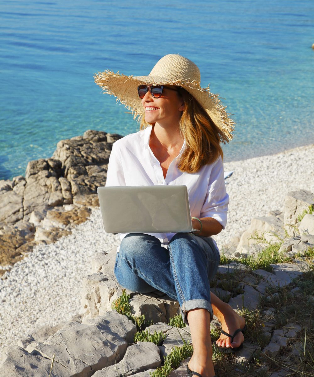 Full length portrait of smiling woman sitting at the rock by the see and using her personal computer. Beautiful female working online. Small business.